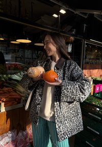 Laget av leopardmønstret, quiltet jakke med store ermer; holder en brun squash og et oransje gresskar. Bakgrunnen viser friske produktskasser.