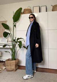 Femme portant un manteau noir, une chemise bleue, un jean et des lunettes de soleil, debout à l'intérieur près d'une grande plante en pot et d'un meuble blanc avec des paniers tressés dessus.