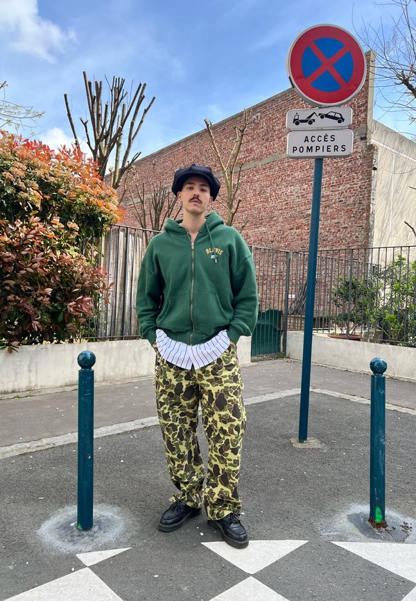 Man met een snor staat op straat naast een parkeerverbodsbord, gekleed in een groene hoodie, camouflagebroek, zwarte schoenen en een marineblauwe pet.