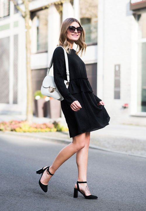 Fekete, bő szárú nadrág, sötétkék, gombos ing fehér szegélyezéssel, piros keresztpántos táska, kék magassarkú cipő, áll a köves utcán.