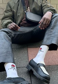 Grüne Bomberjacke, braunes Shirt, schwarze Lederhosen, weiße Socken mit rosemotiv, schwarz-weiße Slipper mit dicker Sohle.