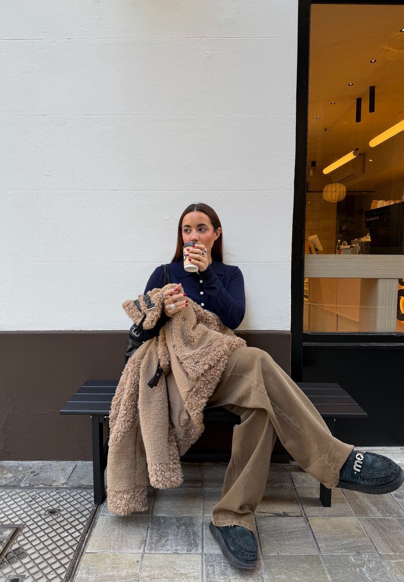 Femme assise sur un banc devant un café, tenant une tasse de café et une veste polaire beige, portant un pantalon marron et des chaussures noires.