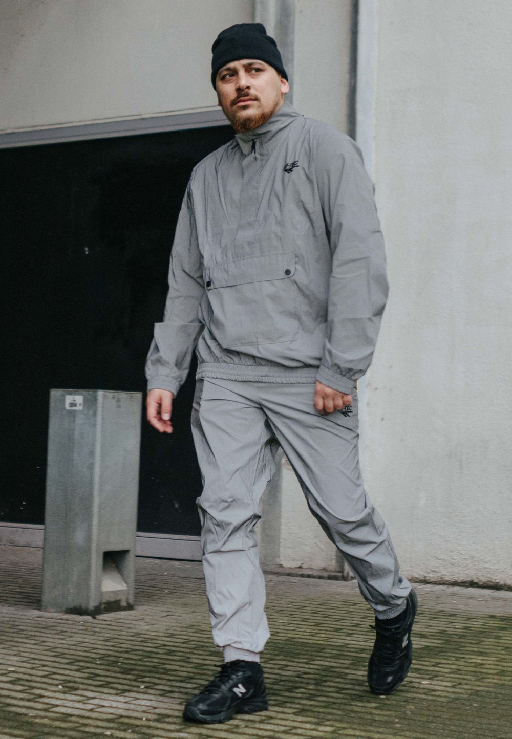 Jasnoszary wiatrówka z zamkiem błyskawicznym, noszona z pasującymi spodniami. Czarne sneakersy, stojąc na fakturowanym chodniku przy ścianie.
