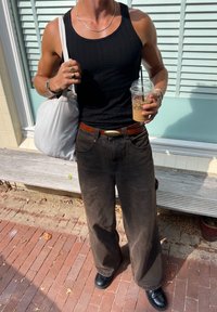 Svart ribbet tanktop, brune vide bukser med belte, sølvring, holder en drink i en mønstret kopp, og en grå tote bag.