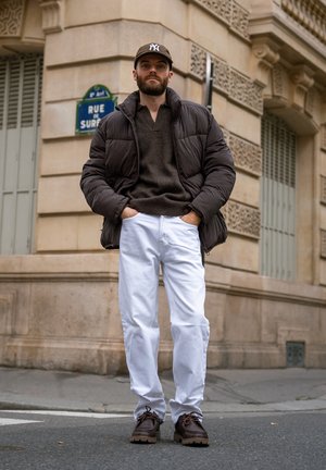 Hombre con barba que lleva una chaqueta acolchada negra, suéter gris, pantalones blancos, zapatos marrones y una gorra de béisbol, de pie con las manos en los bolsillos en una esquina de la calle de la ciudad.
