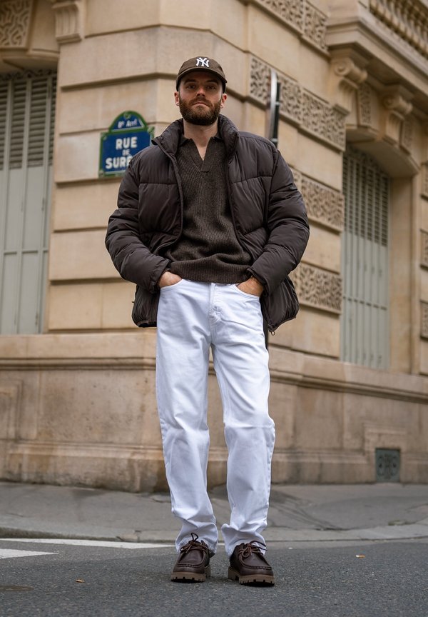 Homme barbu portant une doudoune noire, un pull gris, un pantalon blanc, des chaussures marron et une casquette de baseball, debout les mains dans les poches à un coin de rue en ville.