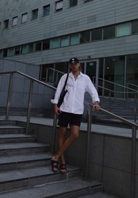 Hvit skjorte med knapper, svarte shorts, svart caps og svarte sandaler. Står på steintrapper med et metallisk rekkverk. Moderne bygning i bakgrunnen.