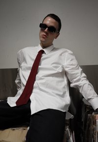 Camisa branca, gravata vermelha, óculos de sol pretos, sentado em caixas. Calças jeans pretas, tecido liso com ligeiro brilho, posicionado de forma casual, fundo neutro.