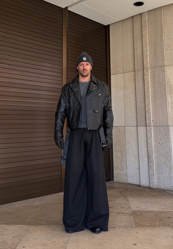 Muž v čiernej koženej bunde, širokých čiernych nohaviciach, sivej mikine, čiernych rukaviciach a čiernej čiapke stojí pred drevenou a kamennou stenou.