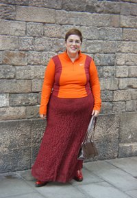 Červená maxi sukňa s textúrou v kombinácii s dlhým opásaným topom v oranžovej a bordovej farbe. Drží hnedú kabelku a nosí červené topánky. Pozadie tvorí kamenná stena.