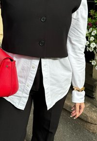 Μαύρο κοντό γιλέκο με δύο κουμπιά πάνω από ένα λευκό υφασμάτινο πουκάμισο, σε συνδυασμό με μαύρο παντελόνι και κόκκινη δερμάτινη τσάντα. Χρυσό ρολόι στο χέρι.