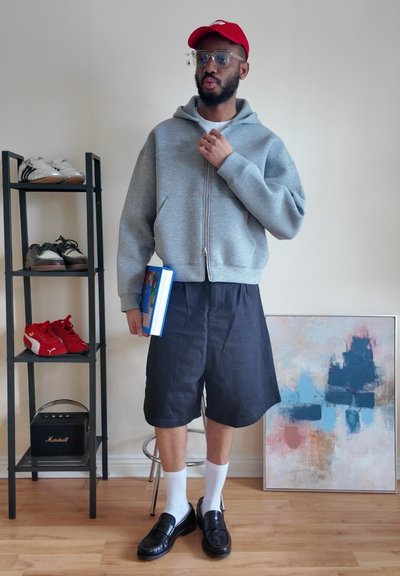 Hombre con gorra roja, sudadera gris con capucha, pantalones cortos negros y mocasines sostiene un libro, de pie cerca de un zapatero y una pintura abstracta sobre un suelo de madera.