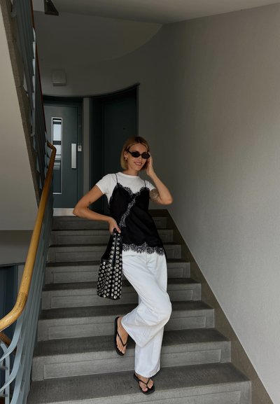 Camiseta de tirantes negra con encaje sobre una camiseta blanca, combinada con pantalones anchos blancos y chanclas negras. Lleva un bolso tote negro con tachuelas.