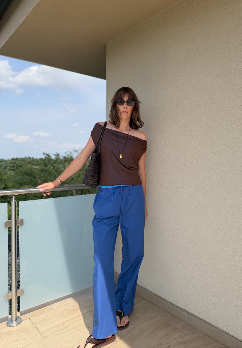 Donna con occhiali da sole, top marrone scollato, pantaloni blu, sandali neri e borsa nera, in piedi su un balcone con muro beige sullo sfondo.