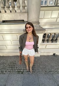 Grijze oversized blazer, roze tanktop, witte kantbroeken, beige kniehoge laarzen, gelaagde ketting met ster- en bloemenaccenten, bruine handtas.