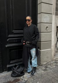 Personne portant un blazer noir surdimensionné, une grande ceinture, un jean bleu clair déchiré, des bottes noires à talons et des lunettes de soleil surdimensionnées, se tenant près d'une porte noire.