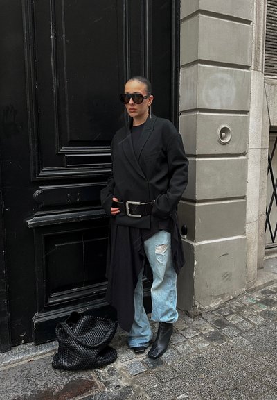 Persona con blazer negro de gran tamaño, cinturón grande, jeans de mezclilla azul claro rasgados, botas negras con tacón y gafas de sol de gran tamaño, de pie junto a una puerta negra.