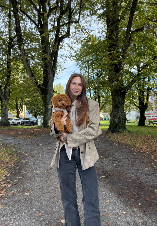 Una mujer con un abrigo beige sostiene a un perro pequeño de color marrón y pelo rizado en un parque frondoso.