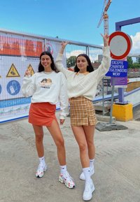 To kvinder poserer udendørs; den ene i en hvid sweatshirt med grafik og en orange miniskirt, den anden i en kremfarvet sweater, en plaid miniskirt og sneakers.
