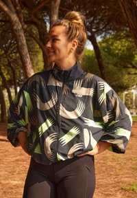 Veste zippée noire avec des motifs abstraits en vert et beige représentant des mains, dotée d'un col haut et de manches larges et amples. Associée à des leggings noirs.