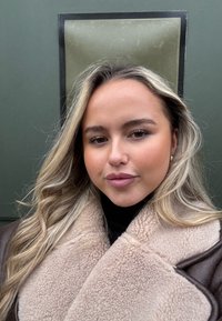 Jeune femme aux longs cheveux blonds, portant une veste en peau retournée beige et un col roulé noir, debout devant un mur vert.