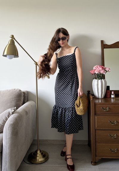 Vestido negro con lunares, con dobladillo acampanado, tirantes finos y detalles de encaje. Un bolso de mimbre marrón y unos tacones burdeos completan el atuendo.