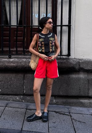 Top negro sin mangas con bordados coloridos, pantalones cortos rojos con rayas blancas, zuecos negros y un bolso de mimbre. De pie sobre un pavimento de piedra.