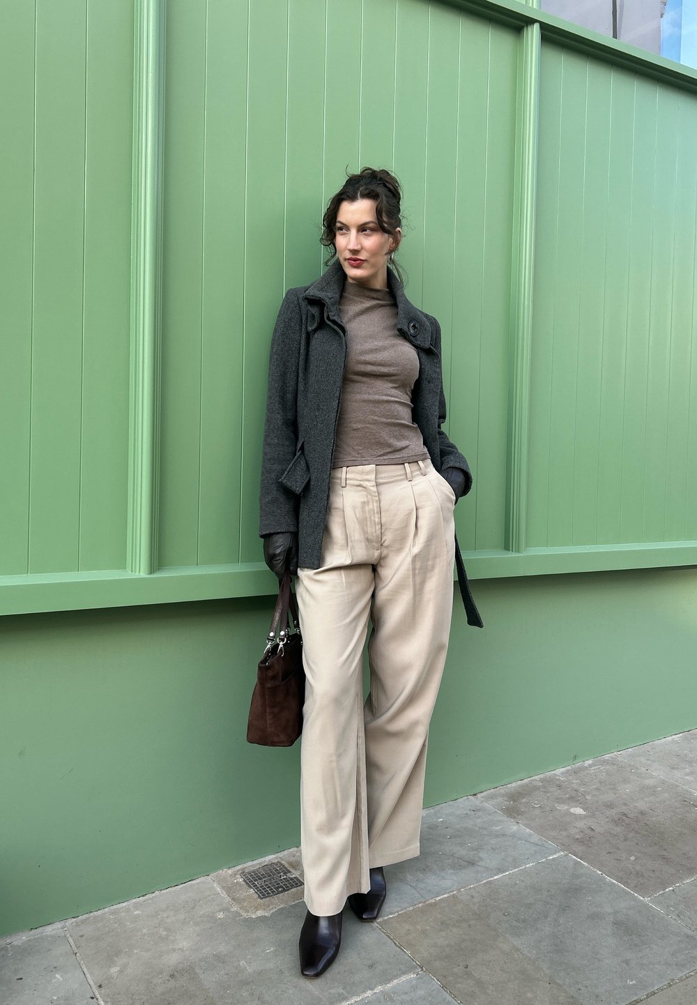 Manteau en laine gris, col roulé marron, pantalon beige, sac à main marron foncé et escarpins noirs pointus devant un mur vert. Textures lisses et coupe ajustée.