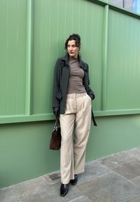 Manteau en laine gris, col roulé marron, pantalon beige, sac à main marron foncé et escarpins noirs pointus devant un mur vert. Textures lisses et coupe ajustée.