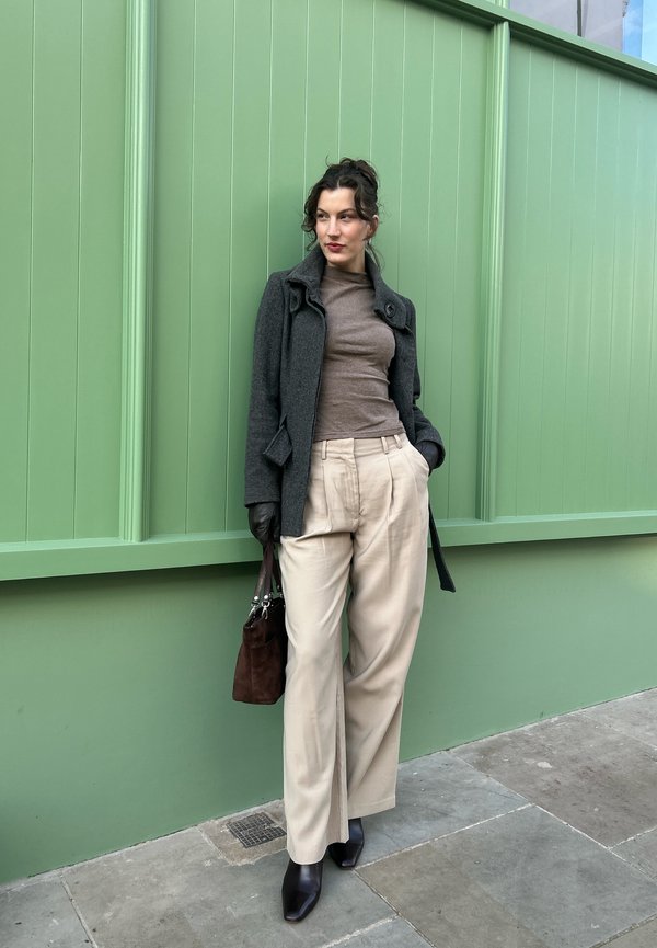 Manteau en laine gris, col roulé marron, pantalon beige, sac à main marron foncé et escarpins noirs pointus devant un mur vert. Textures lisses et coupe ajustée.