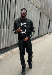 Fekete bőrkabát fekete pulóver felett, amelyen fehér absztrakt minták láthatók, fekete bőr nadrággal és magas szárú fekete csizmával, amelyeken csatok találhatóak.