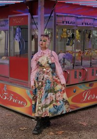 Blumengemustertes Kleid in lebhaften Farben, kombiniert mit einem pinken Hemd und schwarzen Plateau-Stiefeln, vor einem bunten Spielhallen-Hintergrund.