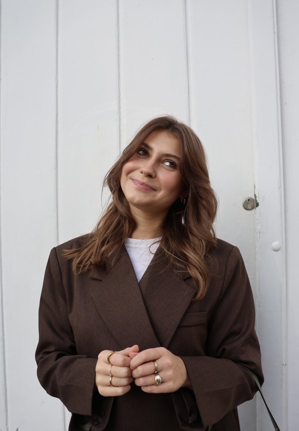 Brun skræddersyet blazer med brede revers, båret over en hvid top. Personen har mellemlangt, bølget hår og holder hænderne sammen.