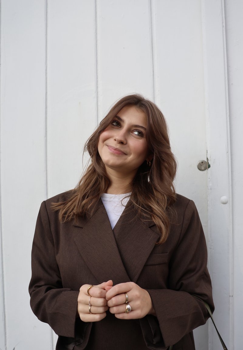 Blazer marron sur mesure avec larges revers, porté sur un haut blanc. Le sujet a les cheveux mi-longs ondulés et tient ses mains jointes.