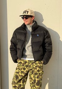 Homme portant une casquette beige, des lunettes de soleil noires, une doudoune noire, un pull gris zippé et un pantalon camouflage jaune-vert, debout devant un mur clair.