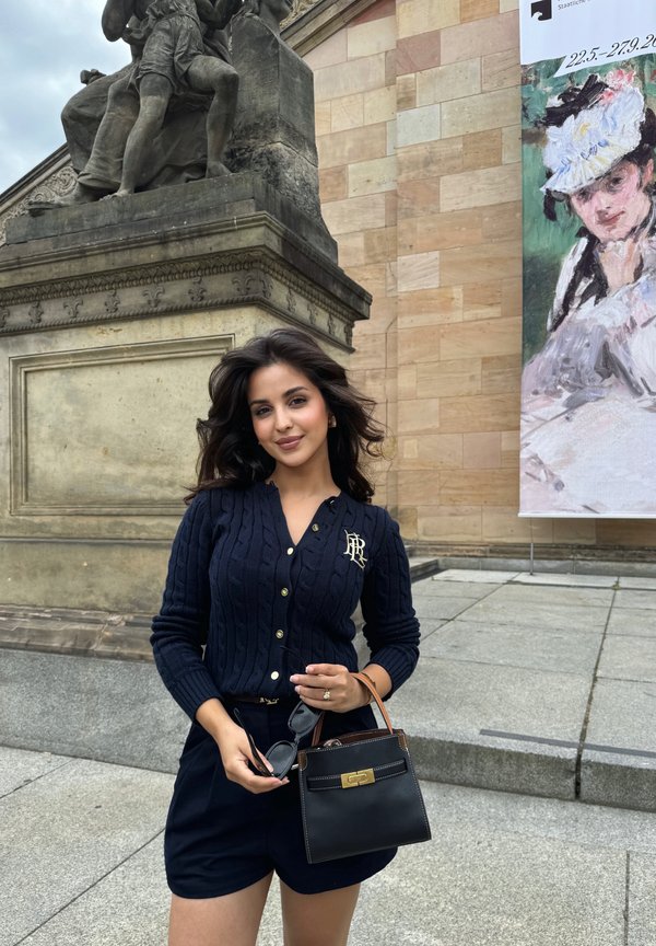 Ung kvinde i navy-outfit, der holder solbriller og sort håndtaske, står nær en statue og et kunstbanner på en stenplads.