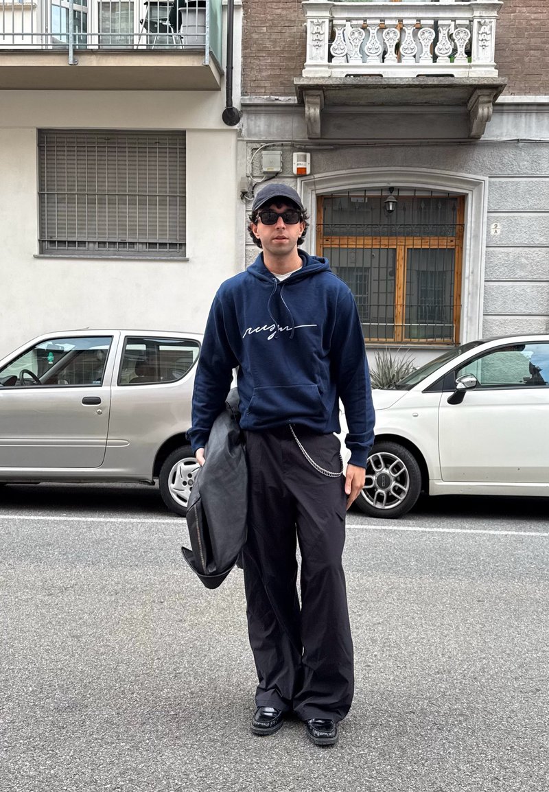 Ναυτικό φούτερ με λευκή γραφή, μαύρο φαρδύ παντελόνι και μαύρα παπούτσια. Τα αξεσουάρ περιλαμβάνουν γυαλιά ηλίου και δερμάτινη τσάντα ώμου. Αστική σκηνή στο φόντο.