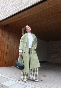 Μακριά, ανοιχτό πράσινη παλτό με μεγάλα κουμπιά, λευκό πουλόβερ και καρό παντελόνι με φαρδύ πόδι. Μαύρη τσάντα με χρυσές λεπτομέρειες. ΚCasual υποδήματα.
