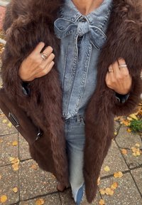 Brązowy futrzany płaszcz z fakturowym wykończeniem, dżinsowa koszula z muszką oraz niebieskie dżinsy. Metalowe pierścionki na dłoniach i brązowa torebka.