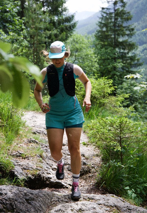 Junge Frau in blauer Sportkleidung und Cap läuft auf einem felsigen Weg, umgeben von dichtem grünem Wald und Bergen im Hintergrund.