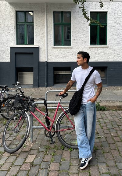Bicicleta rosa con una cesta negra aparcada en un empedrado. Persona vestida con una camiseta blanca, pantalones vaqueros anchos de color azul claro y un bolso negro texturizado al hombro.