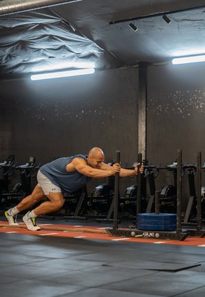 A person pushes a weight sled on a rubber floor in a gym. The sled has blue weights and is positioned near rowing machines.