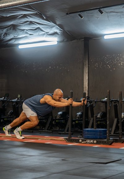 Una persona empuja un trineo de pesas sobre un suelo de goma en un gimnasio. El trineo tiene pesas azules y está ubicado cerca de las máquinas de remo.