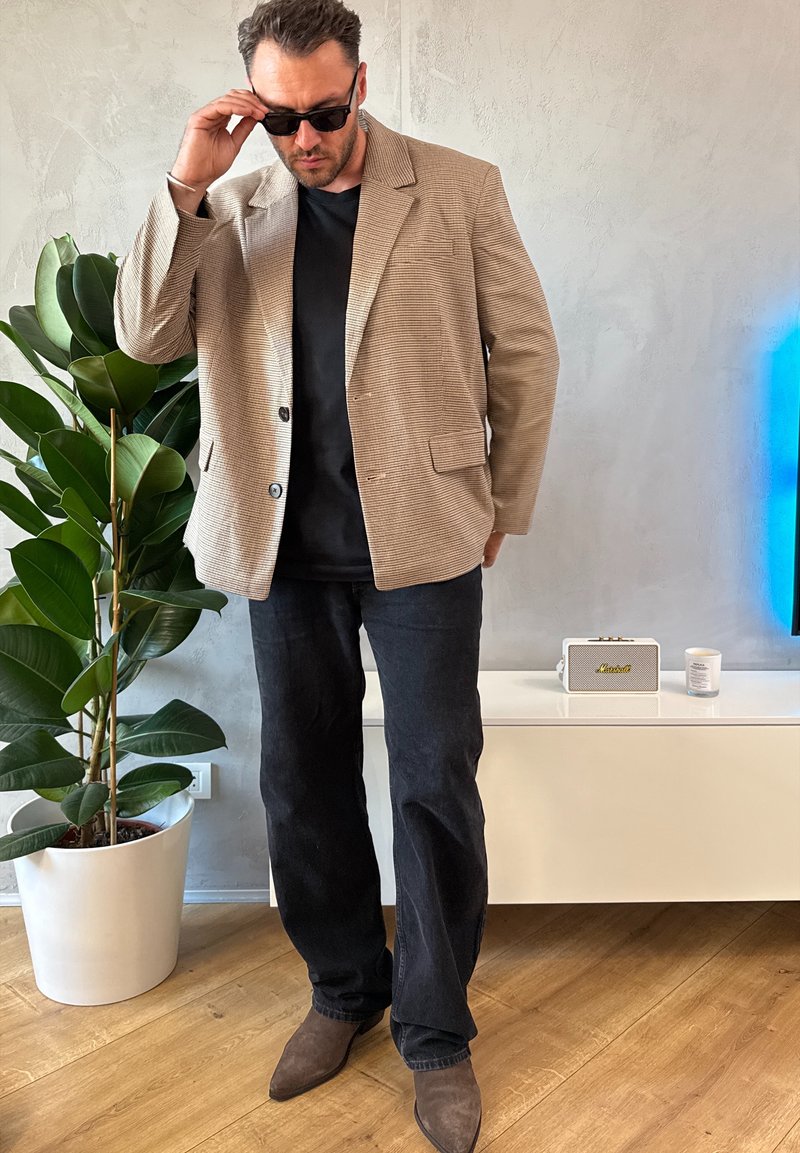 Braun kariertes Sakko über einem schwarzen Hemd, kombiniert mit dunklen Jeans und braunen Stiefeletten. Im Hintergrund sind eine Pflanze und ein kleiner Lautsprecher zu sehen.