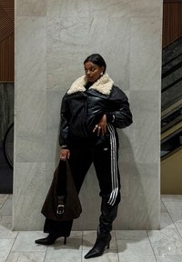 Kvinde iført sort læderjakke med hvid pelskrave, sorte Adidas træningsbukser, sorte støvler med hæl, holder en stor brun taske, står indendørs.