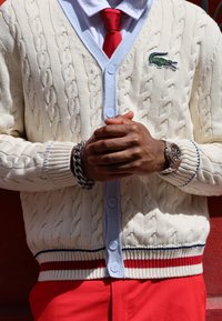 Πλεκτό σακάκι από ελεφαντόδοντο με σχέδιο καλωδίου, ανοιχτό μπλε γιακά και κόκκινο και μπλε ριγέ στρίφωμα. Περιλαμβάνει πράσινο κεντημένο λογότυπο Lacoste.