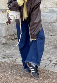 Brown leather bomber jacket, yellow sweater, wide blue denim jeans, black sneakers with white stripes, and a white handbag with a charm.