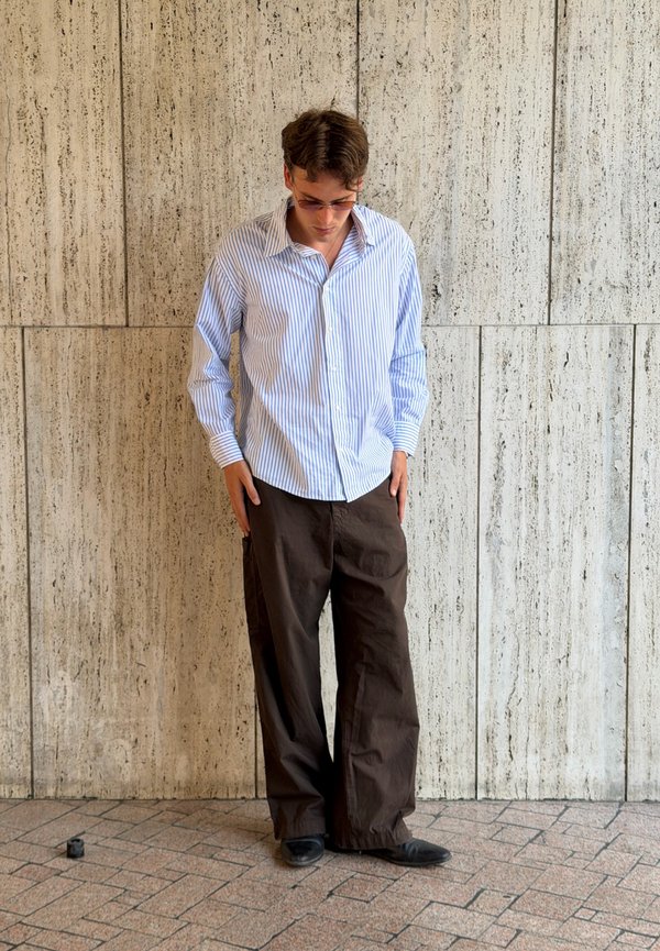 Jeune homme portant une chemise bleue rayée, un pantalon large marron et des chaussures noires, debout en regardant vers le bas sur un fond de mur en pierre beige.