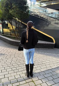 Crna skraćena jakna, svijetloplave uske traperice i crne čizme do koljena; kosa u pletenici, stoji ispred modernog urbanog okruženja.