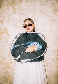 Casaco preto com fecho éclair e riscas brancas, saia branca e mala azul oversized. Modelo usa óculos de sol castanhos e joias em camadas.
